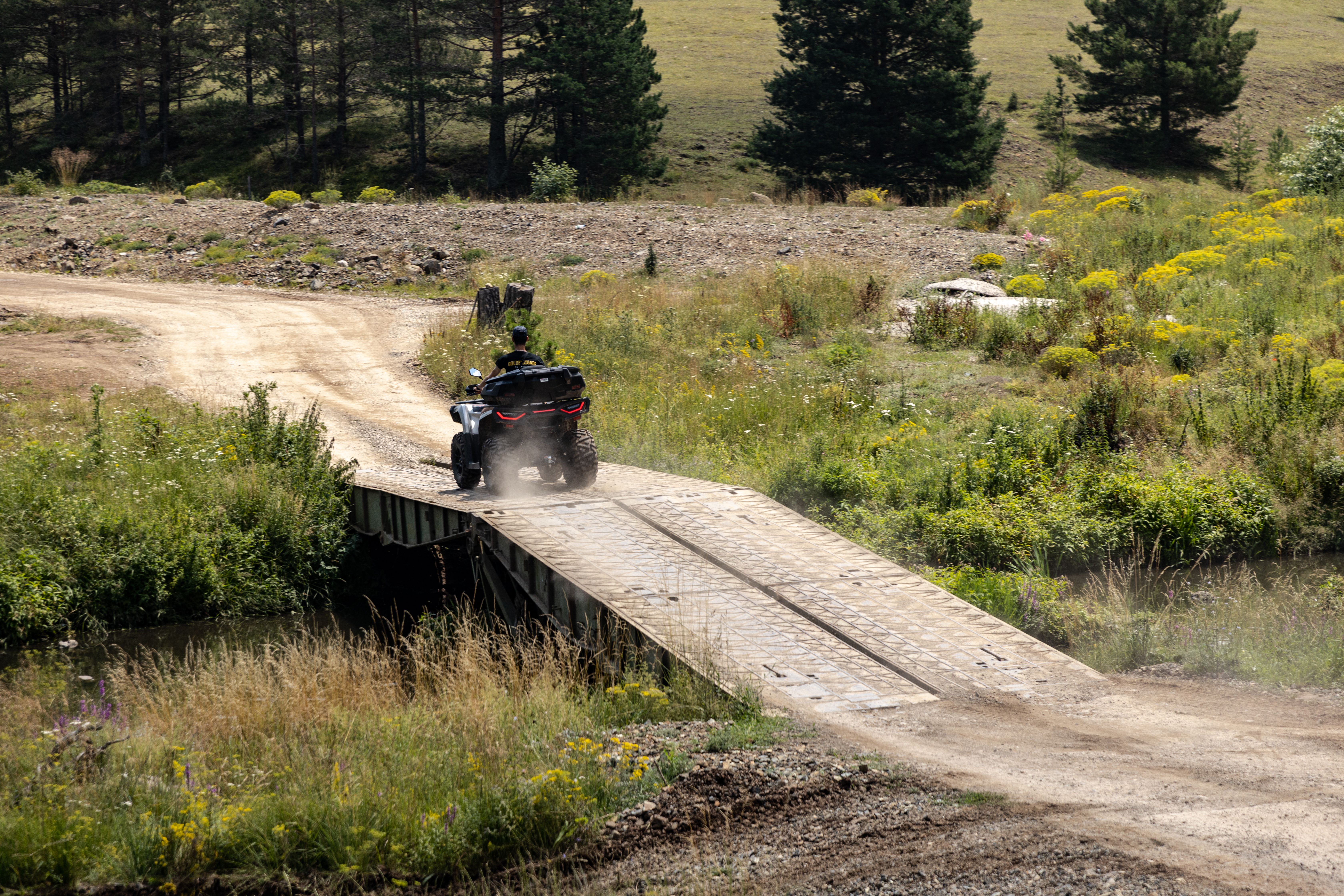 An ATV crosses a rustic bridge surrounded by lush greenery in the sunny Zlatibor region of Serbia.