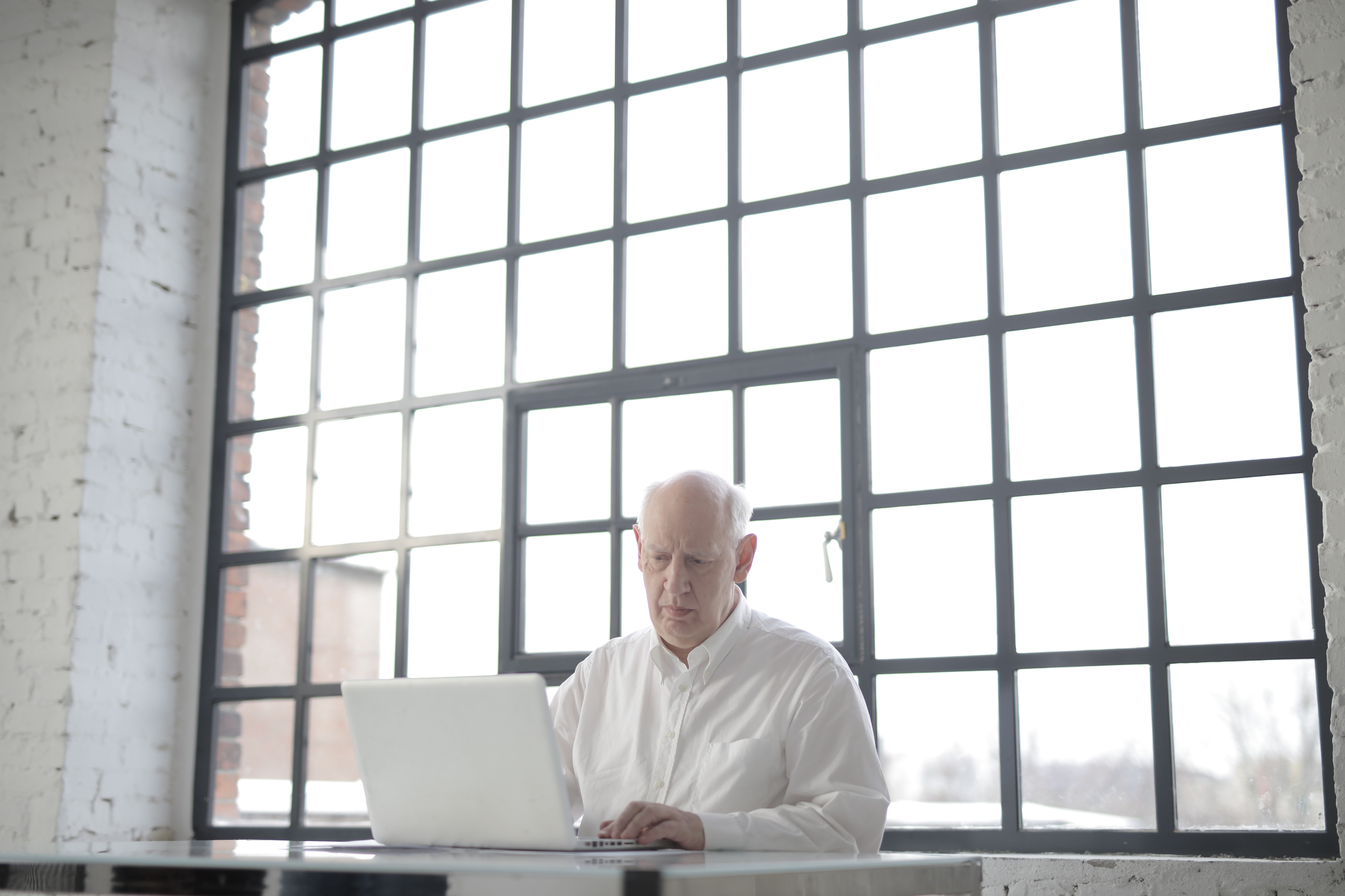 A senior man working on a laptop in a bright office with large windows.