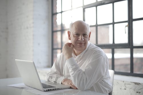 Positive bold aged male freelancer wearing formal shirt sitting at table with laptop and documents and looking at camera with smile during remote work in room with industrial interior