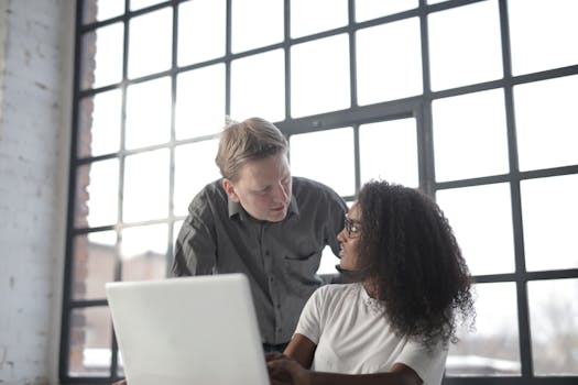 Two colleagues engaged in a business discussion in a bright, modern office with large windows.