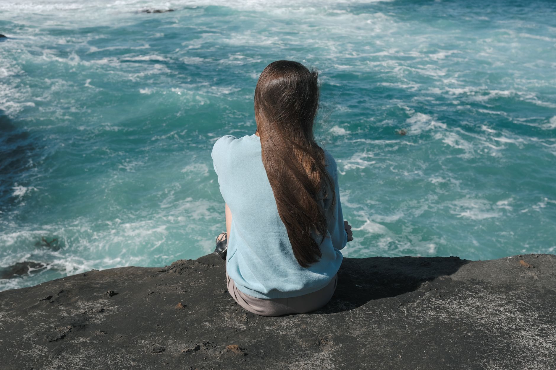 A woman with long hair sits on a cliff edge, enjoying a windy ocean view in Victoria, Australia.