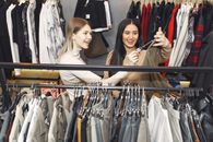 From above of young cheerful friends choosing casual pullover and laughing in shop while female looking at tag of wear