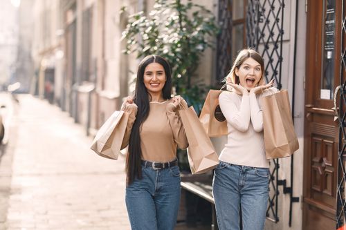 Free Positive young casually dressed women with shopping bags standing near building while smiling and looking at camera in sunny summer day Stock Photo