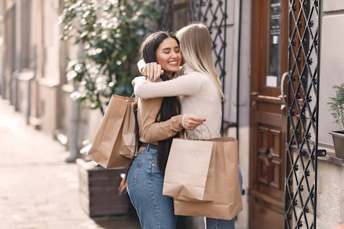 Side view of cheerful young women with shopping bags standing on city street near door while toothy smiling and hugging