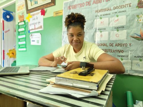 Free Smiling teacher sitting at desk in a vibrant classroom, surrounded by educational materials and documents. Stock Photo