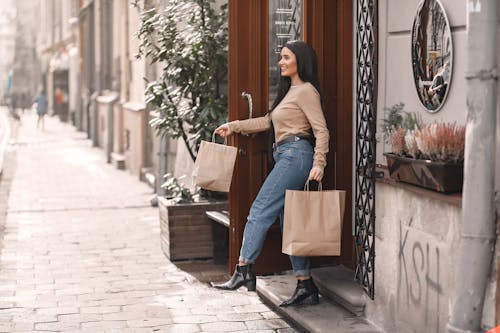 Free Side view of happy dark haired woman with paper shopping bags stepping on stairs while coming out of shop building in sunny day Stock Photo