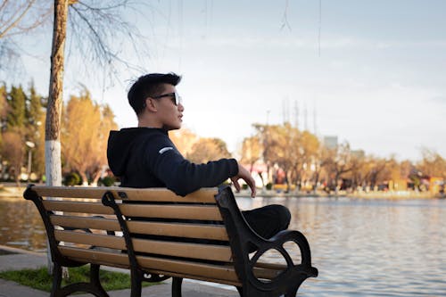 坐在水边附近的长椅上，戴着眼镜的黑夹克的男人