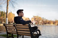 Man in Black Jacket Wearing Eye Glasses Sitting on Bench Near Body of Water