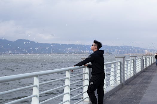 一个男人穿着休闲服，站在码头栏杆上，凝视着大海和远山