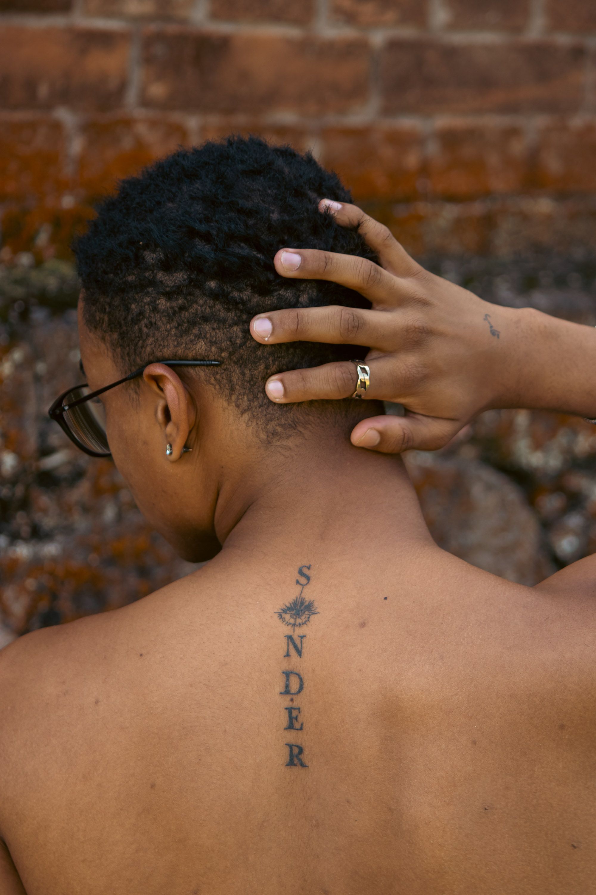 A person shows a 'Sonder' tattoo on their back, hand touching head, against a brick wall.