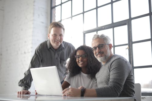 Free Optimistic diverse managers in casual wear smiling at camera while using computer and discussing details of work in light office Stock Photo