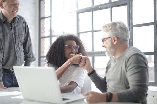 Free Happy colleagues of different ages wearing informal wear and celebrating successful end of project and bumping with fists while using laptop in modern workspace Stock Photo