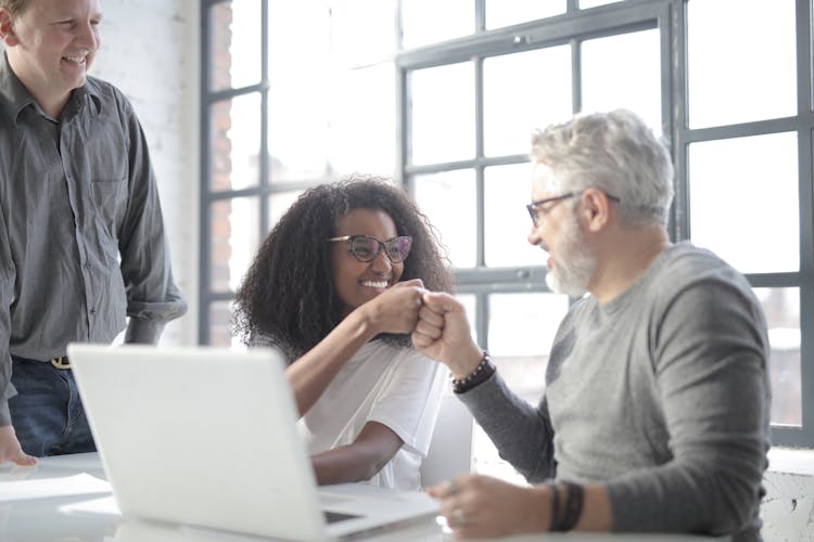 Cheerful Multiethnic Coworkers Greeting Each Other With Fists Surfing Computer In Office