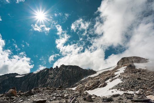 Rugged mountain terrain with snow patches under a bright sun and vibrant blue sky.