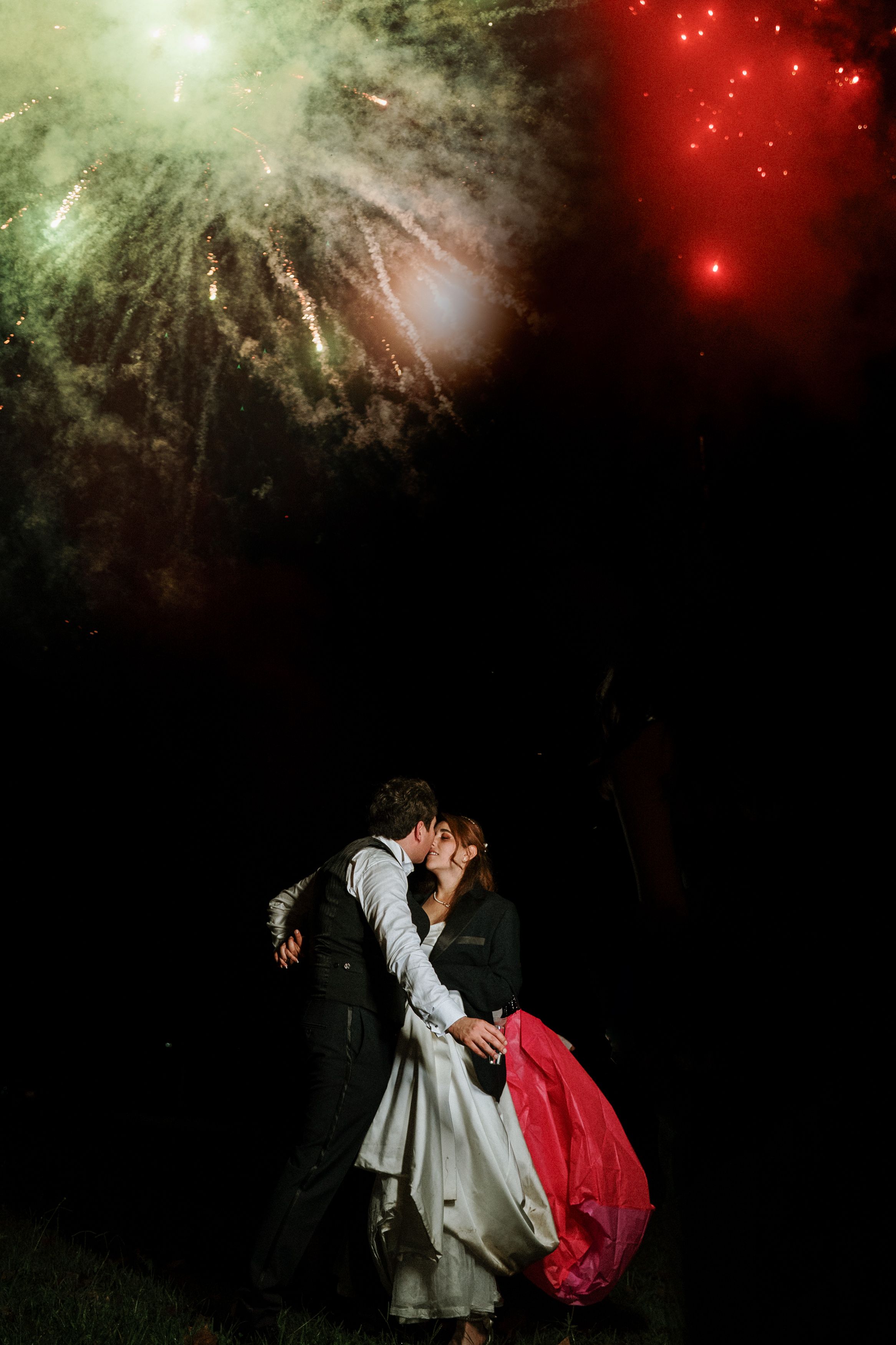 A couple shares a romantic kiss beneath a night sky illuminated by vibrant fireworks.