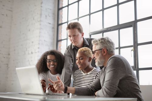 Free Group of multiracial colleagues laughing and pointing to screen of laptop while sitting at table and working on project in modern workplace Stock Photo