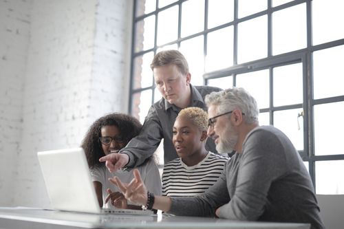 Free Cheerful diverse colleagues working together while man in gray shirt pointing to screen of netbook and old male in glasses gesticulating when explaining project Stock Photo