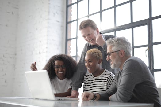 Four colleagues joyfully collaborating on a project with a laptop in a bright modern office.