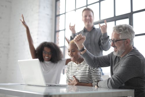 Happy diverse people raising hands and joyfully laughing while looking at screen of laptop and sitting at table in modern loft styled workspace