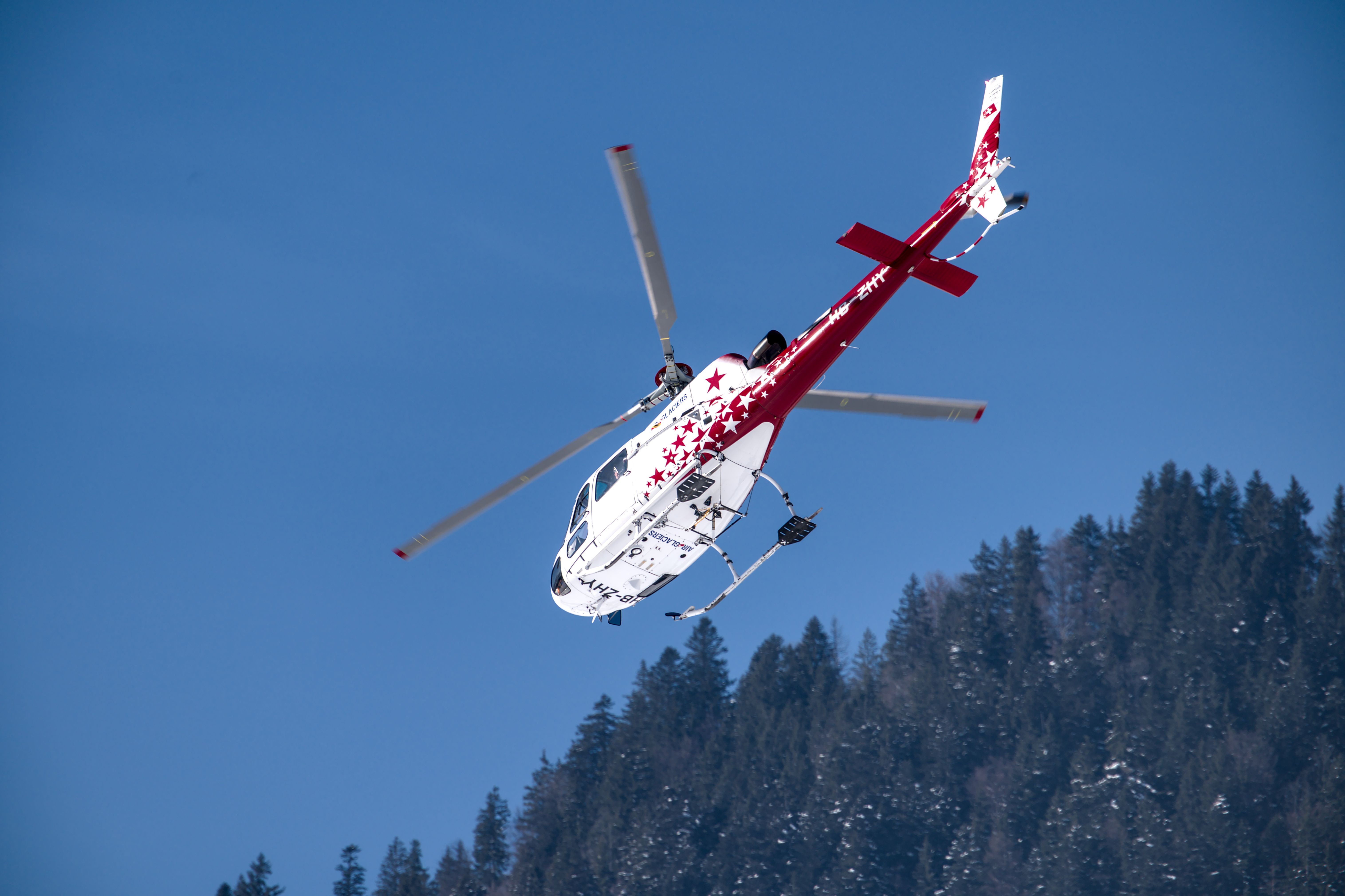 A vibrant red and white helicopter flying above a snowy forest landscape on a clear day.