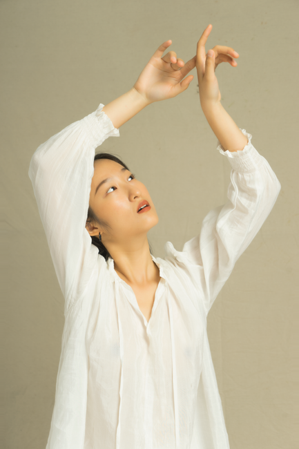 Portrait of an Asian woman in a white blouse, posing elegantly with arms raised, against a neutral background.