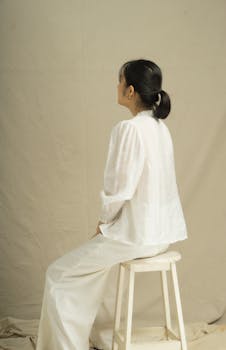 Elegant minimalist portrait of an Asian woman sitting on a stool in a studio setting.