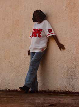 A woman in casual attire leans stylishly against a textured wall in Zaria, Nigeria.