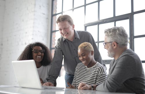 Group of diverse people laughing and using netbook while working on project together in office with white brick walls and big window