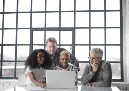 Free Focused serious multiracial colleagues pensively looking at laptop while working on project together and sitting at white table against big window Stock Photo