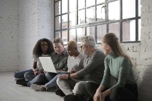 Concentrated serious diverse coworkers surfing laptop and working on project while sitting on floor and leaning on white brick wall