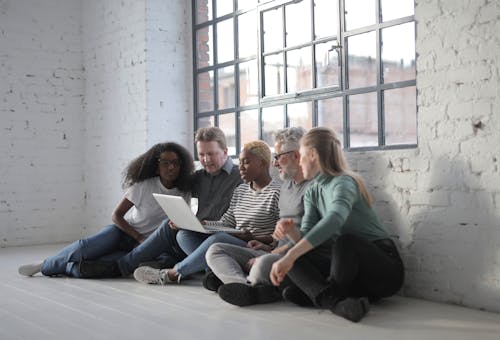 Focused serious multiethnic friends surfing internet on netbook while resting on floor of loft styled workspace with brick walls and big window