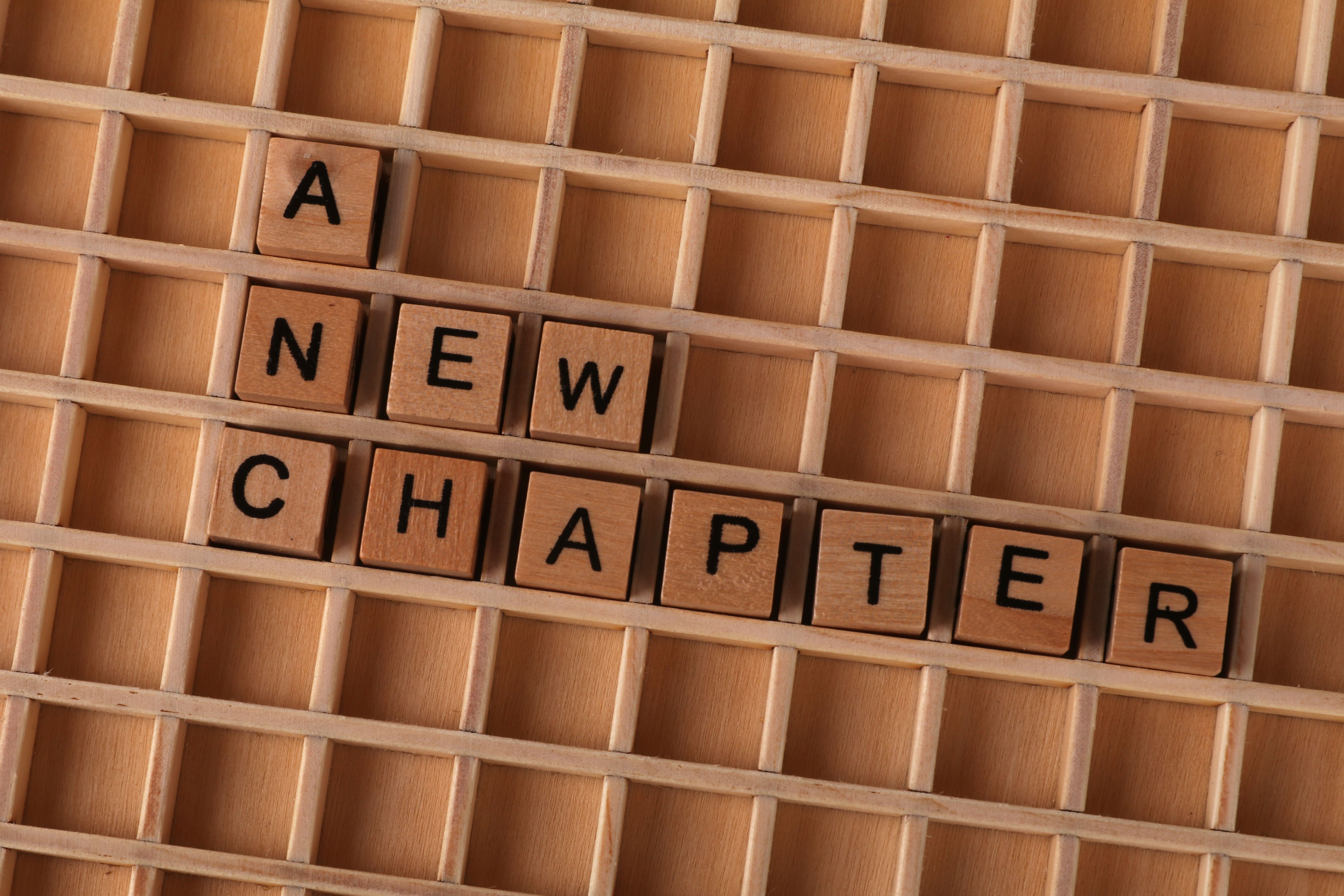 Scrabble tiles spelling 'A New Chapter' on wooden grid, symbolizing new beginnings.