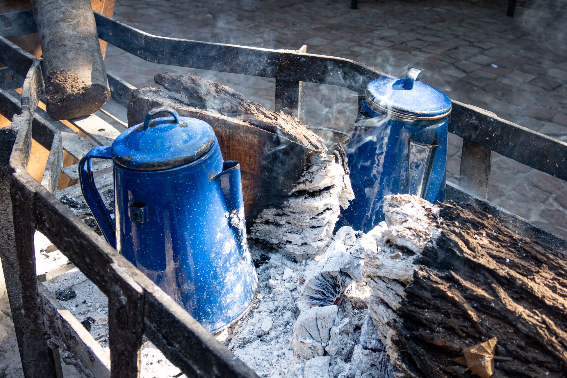 Outdoor campfire scene with blue coffee pots brewing, surrounded by wood and ashes.