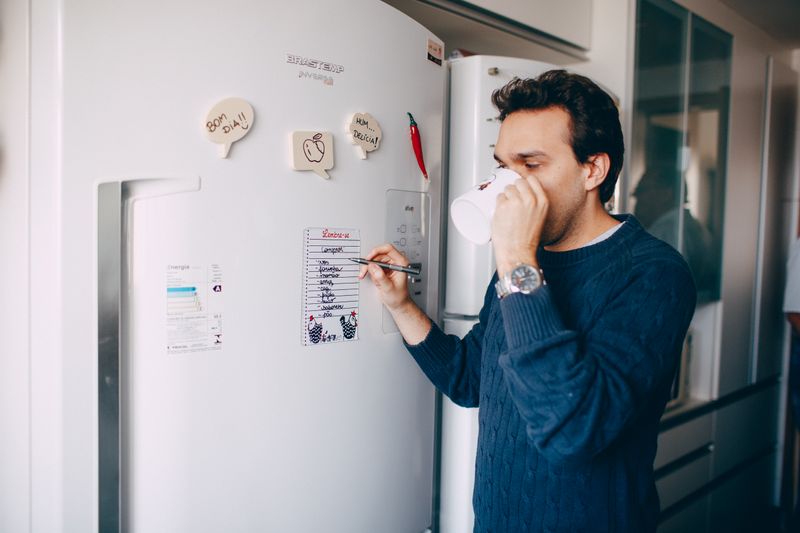 Young male employee in casual outfit drinking coffee and taking notes on paper attached on fridge in morning