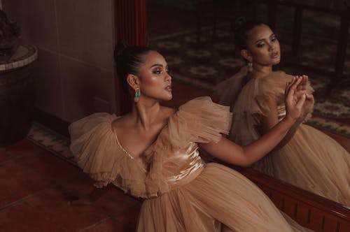 High angle of gorgeous young ethnic female in  elegant gown sitting on floor and looking in mirror over shoulder