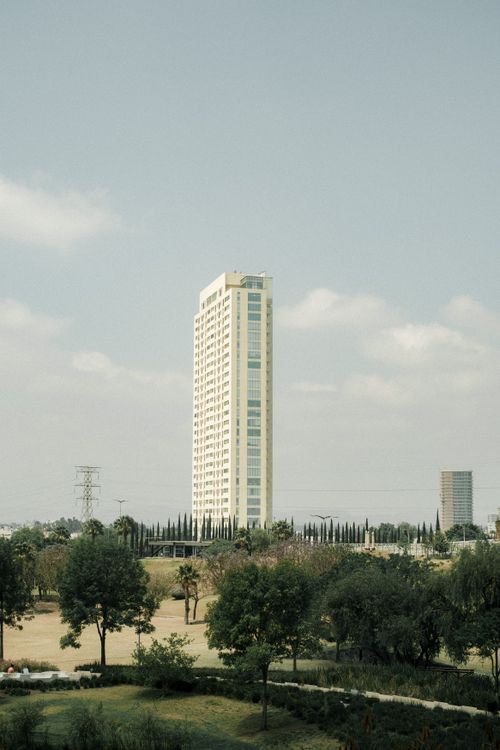 Free A modern skyscraper amidst an urban park, with greenery and a cloudy sky backdrop. Stock Photo