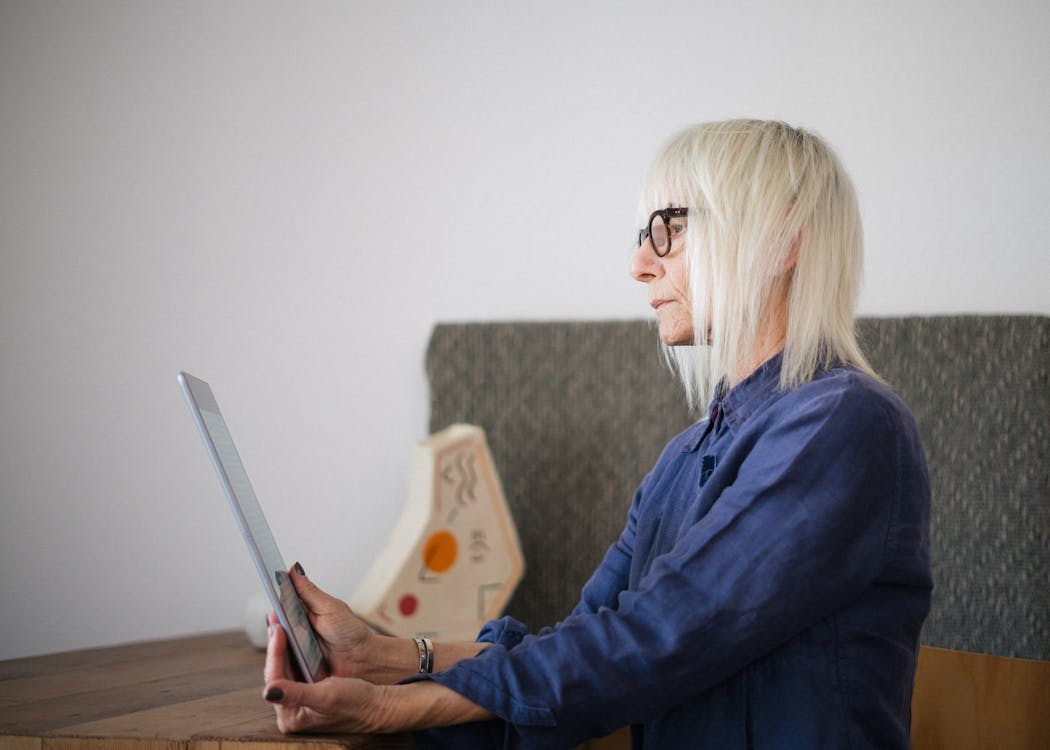 Free Side view of stylish concentrated aged woman in casual outfit sitting with tablet at table at home Stock Photo