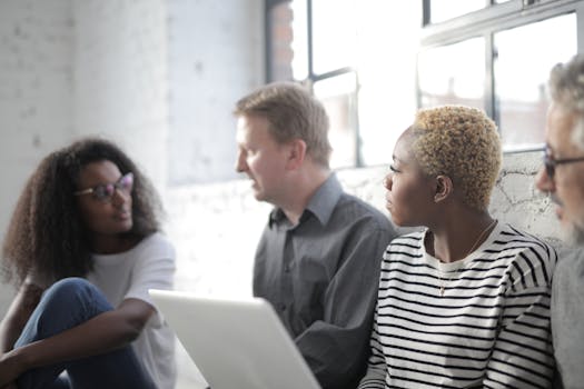 A diverse group of adults engaging in a casual meeting by a window with laptops.