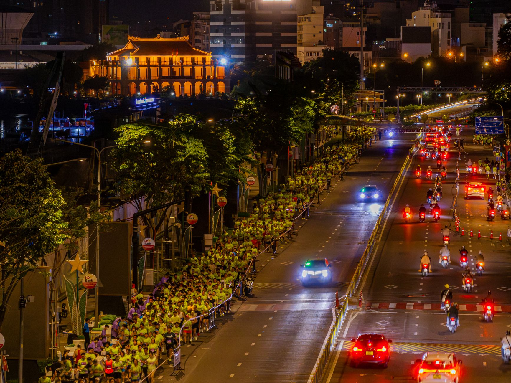 Night marathon with participants and vehicles in a lively city street under illuminated skyline.