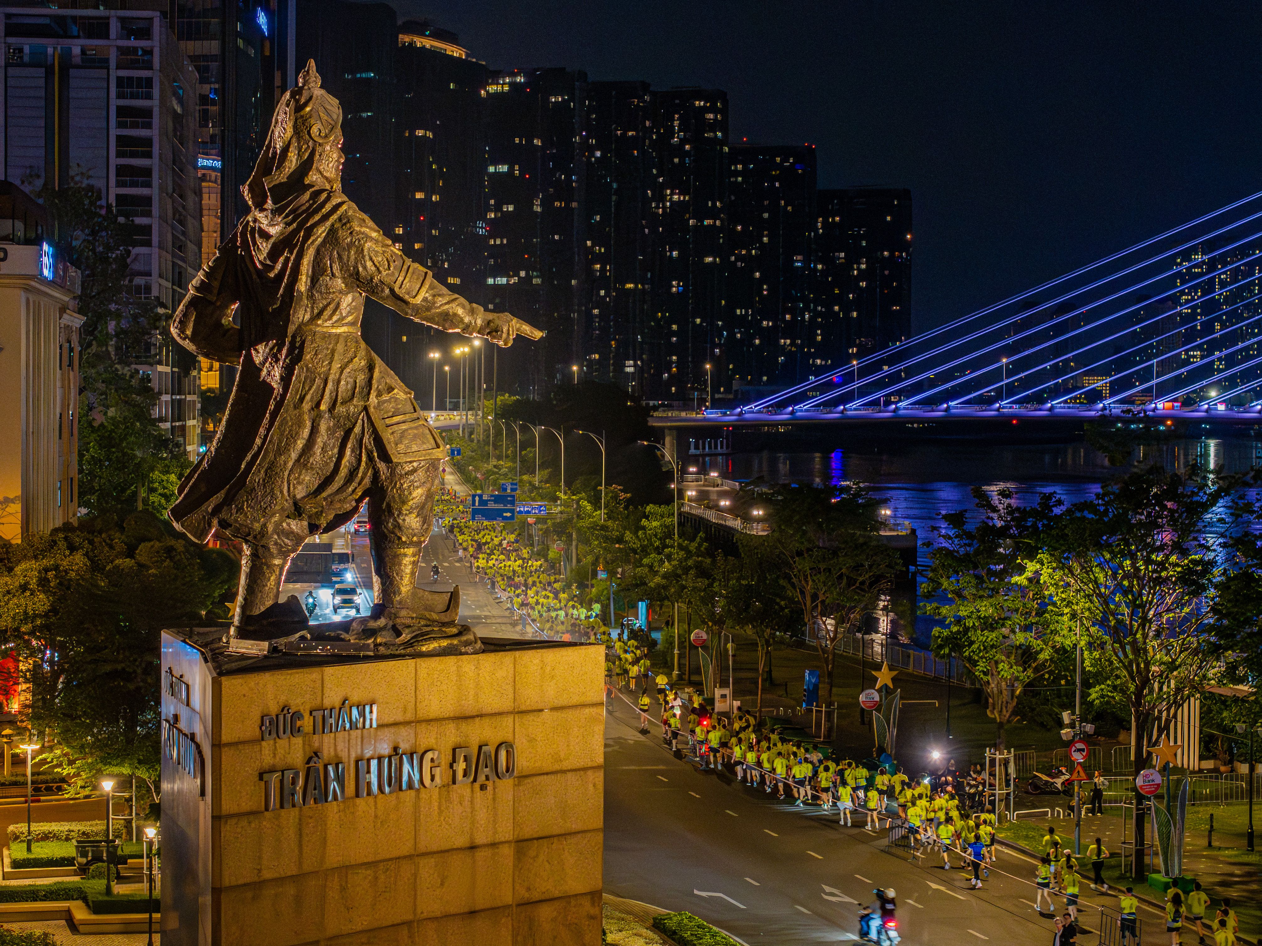 Bustling city night scene with Tran Hung Dao statue and illuminated bridge.