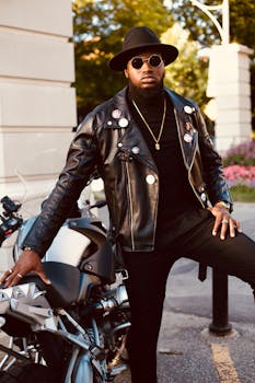 一位神情严肃、留着胡须的非裔美国年轻男子，身穿全套黑色衣帽，戴着太阳镜，站在现代摩托车旁，面向绿色公园