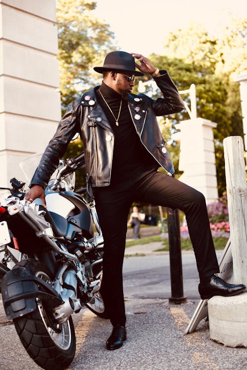 Free Full length of young stylish black male in trendy leather jacket and hat relaxing on street near parked modern motorcycle Stock Photo