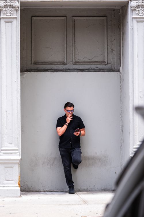 Free Man in black attire leans against building exterior, New York City, smoking and using smartphone. Stock Photo