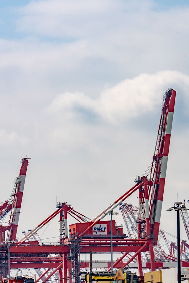 Red and white cargo cranes dominate the skyline in a busy industrial port under a cloudy sky.