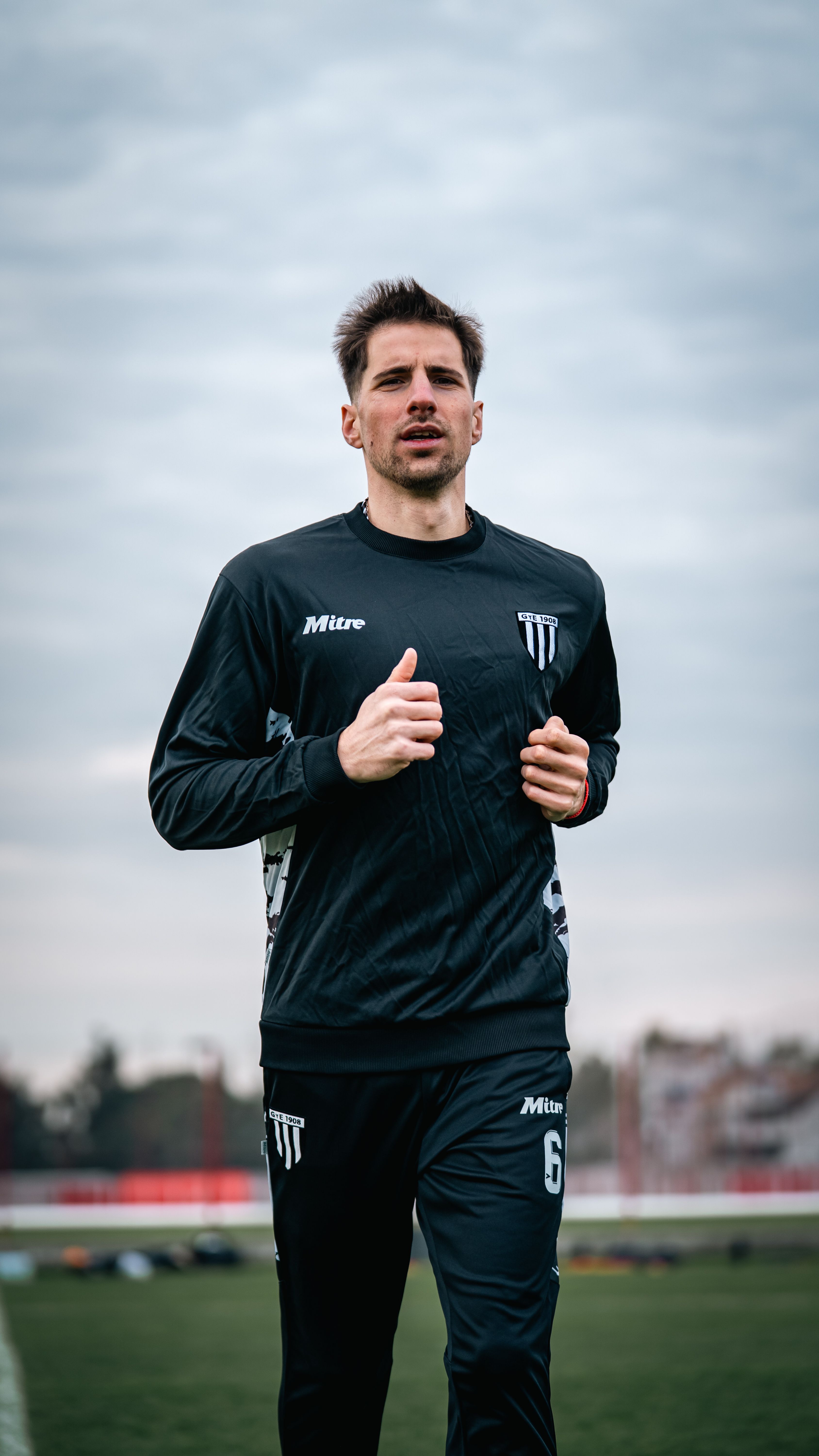 Young male athlete in black sportswear jogging on an outdoor track under a cloudy sky.