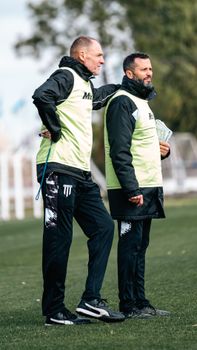 Two soccer coaches in discussion during outdoor training, wearing team vests.