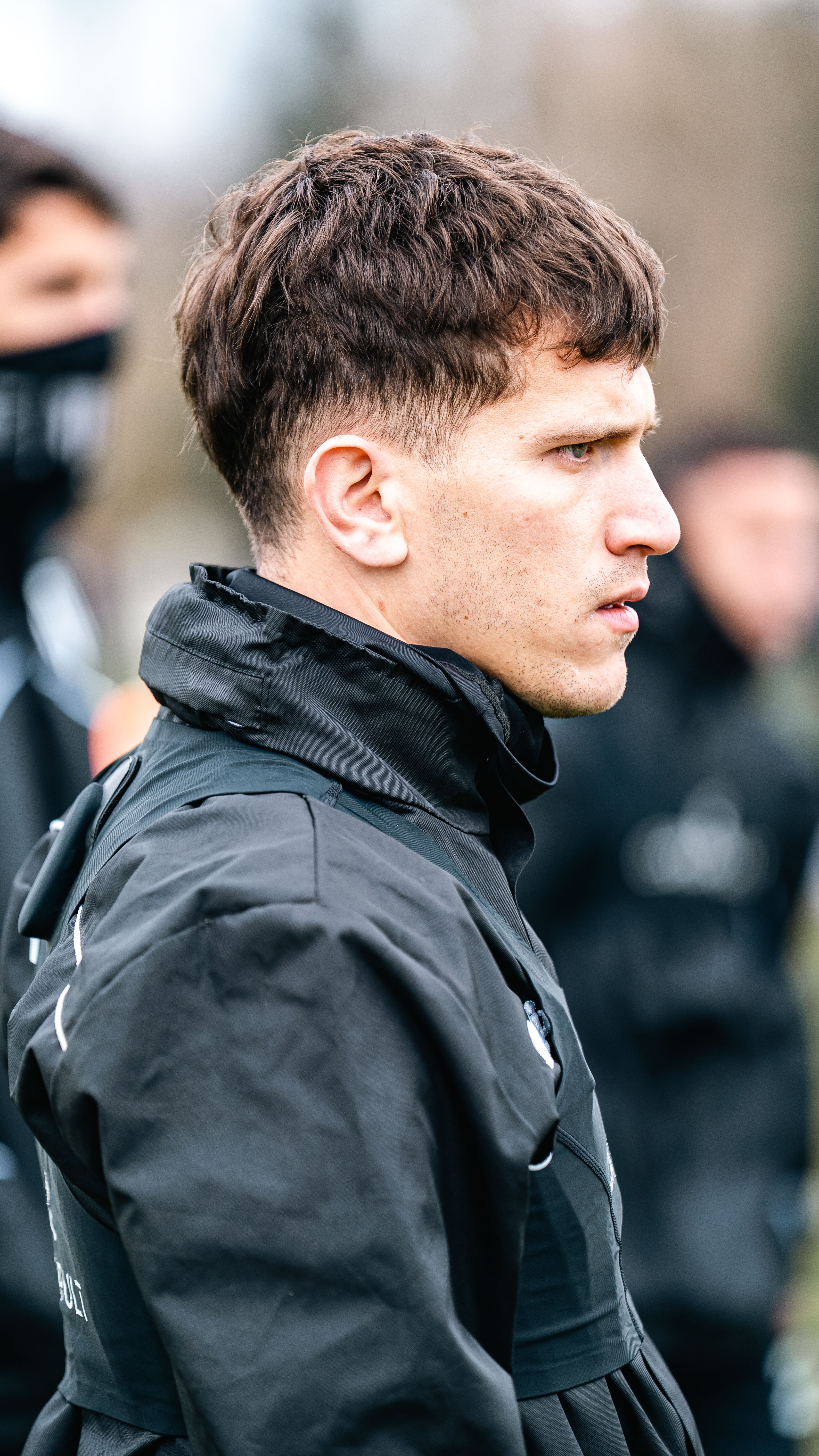 Side profile of a young man wearing a black jacket, deep in thought outdoors.