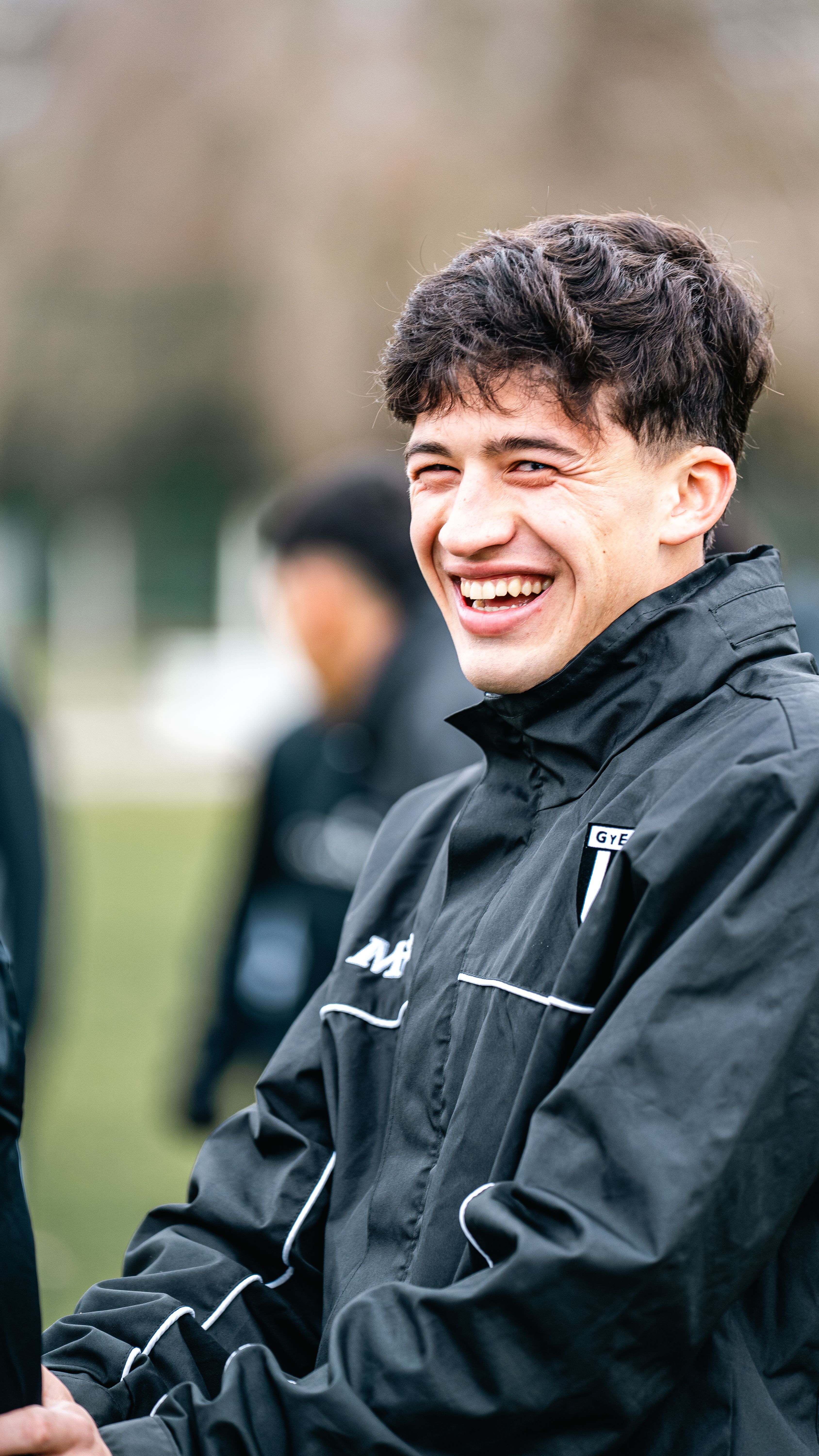 A young man smiles joyfully while wearing a sports jacket outdoors.