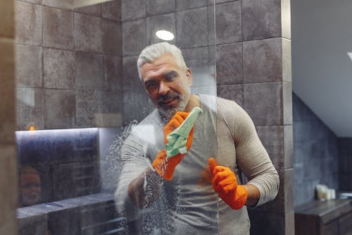 Grey haired male with beard in rubber gloves cheerfully cleaning shower glass shield