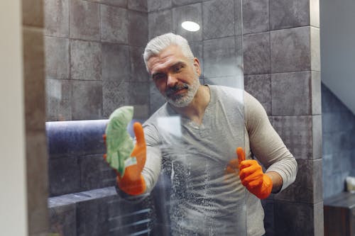 Grey haired male with beard in orange rubber gloves attentively cleaning shower glass in bathroom